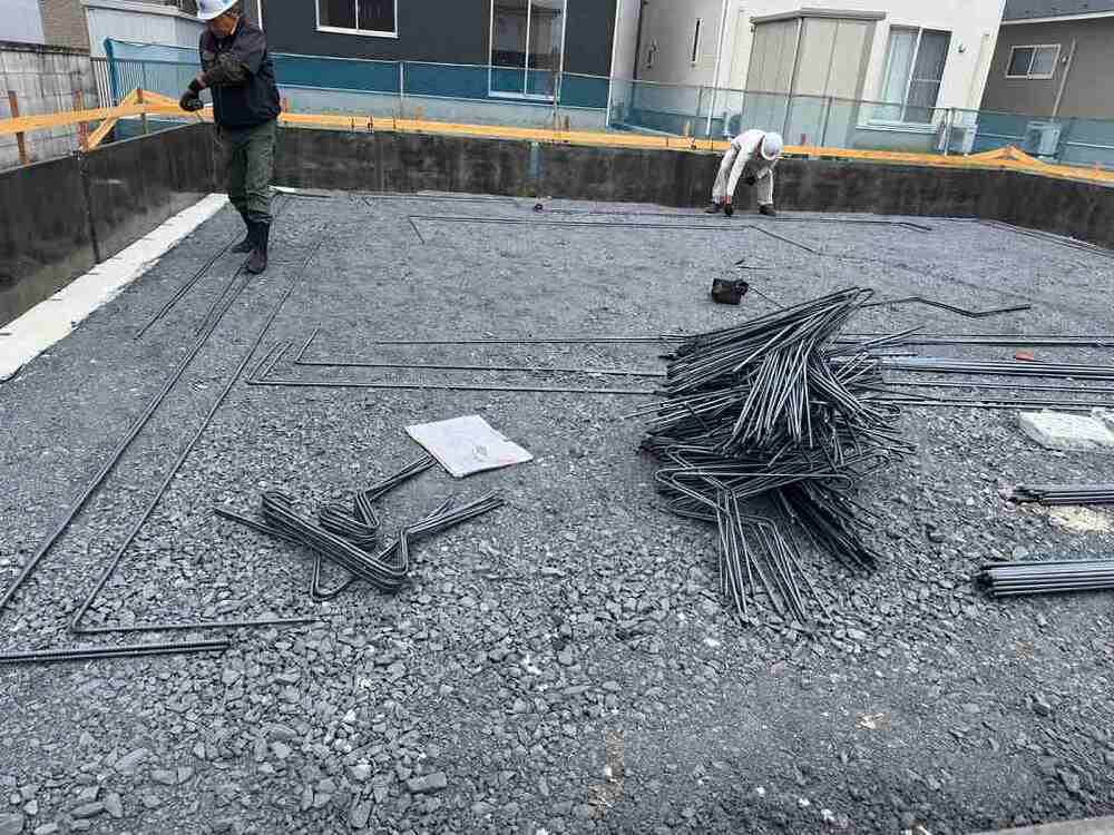 基礎配筋工事の現場で、砕石の上に鉄筋が並べられ、職人が基礎の鉄筋を配置している画像
